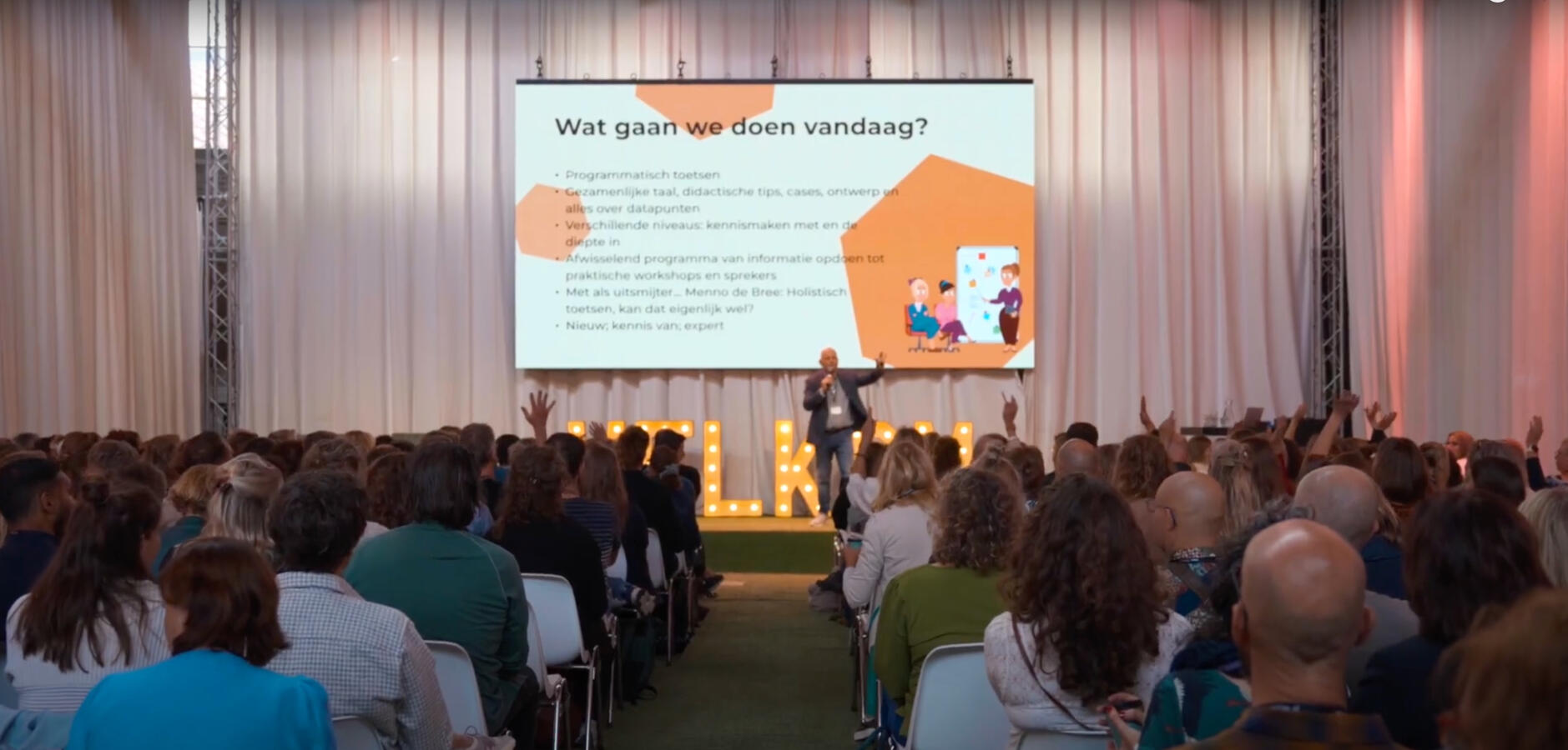 Een volle congreszaal, met een spreker op het podium en deelnemers die hun hand opsteken